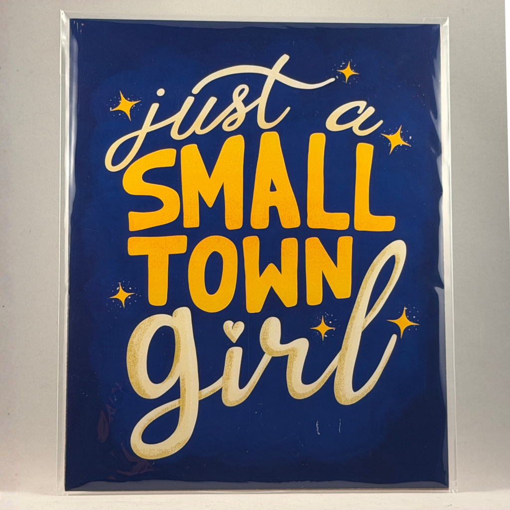A 8x10 art print with the phrase 'just a SMALL TOWN girl' in white and yellow lettering on a dark blue background, photographed in a clear plastic sleeve against a light gray wall.