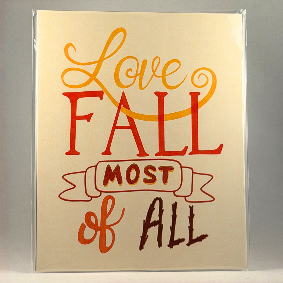 A 8x10 art print featuring the phrase 'Love Fall Most of All' in stylized autumn colors, presented in a clear plastic sleeve against a neutral gray background.