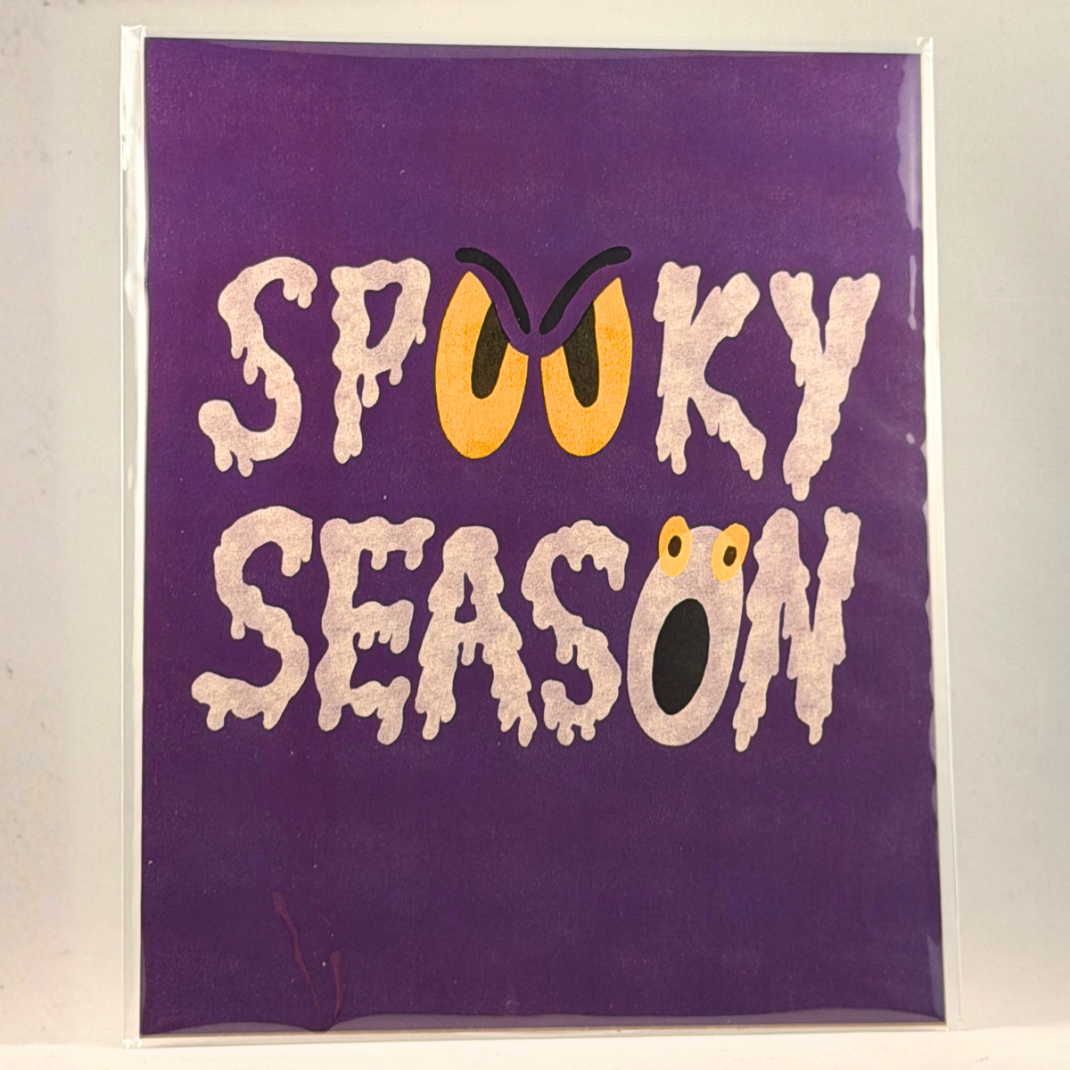A 8x10 purple art print featuring the words 'SPOOKY SEASON' in dripping white letters with Halloween-themed eye and mouth details, photographed against a light background.