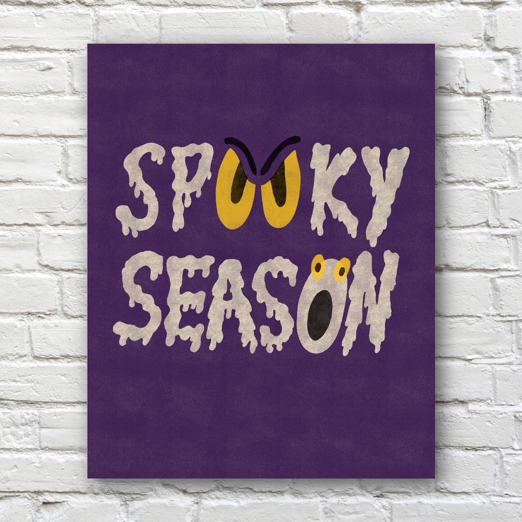 A vertical purple wall art print with the words 'Spooky Season' in dripping white lettering, featuring yellow eyes and a frog face detail, displayed against a white brick wall.