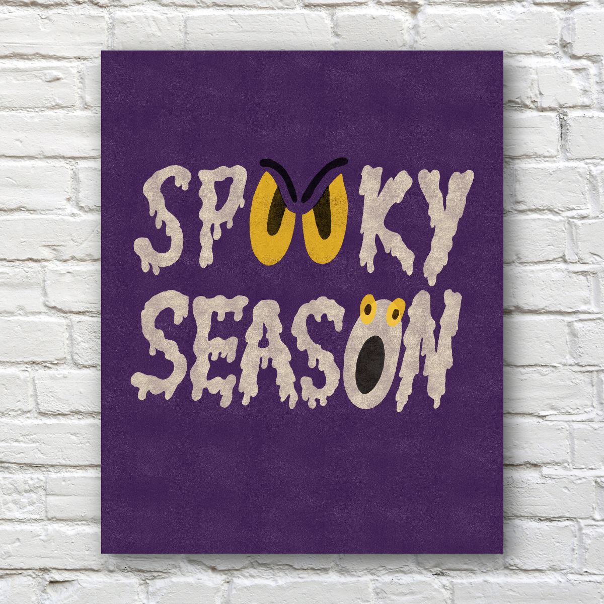 A vertical purple wall art print with the words 'Spooky Season' in dripping white lettering, featuring yellow eyes and a frog face detail, displayed against a white brick wall.
