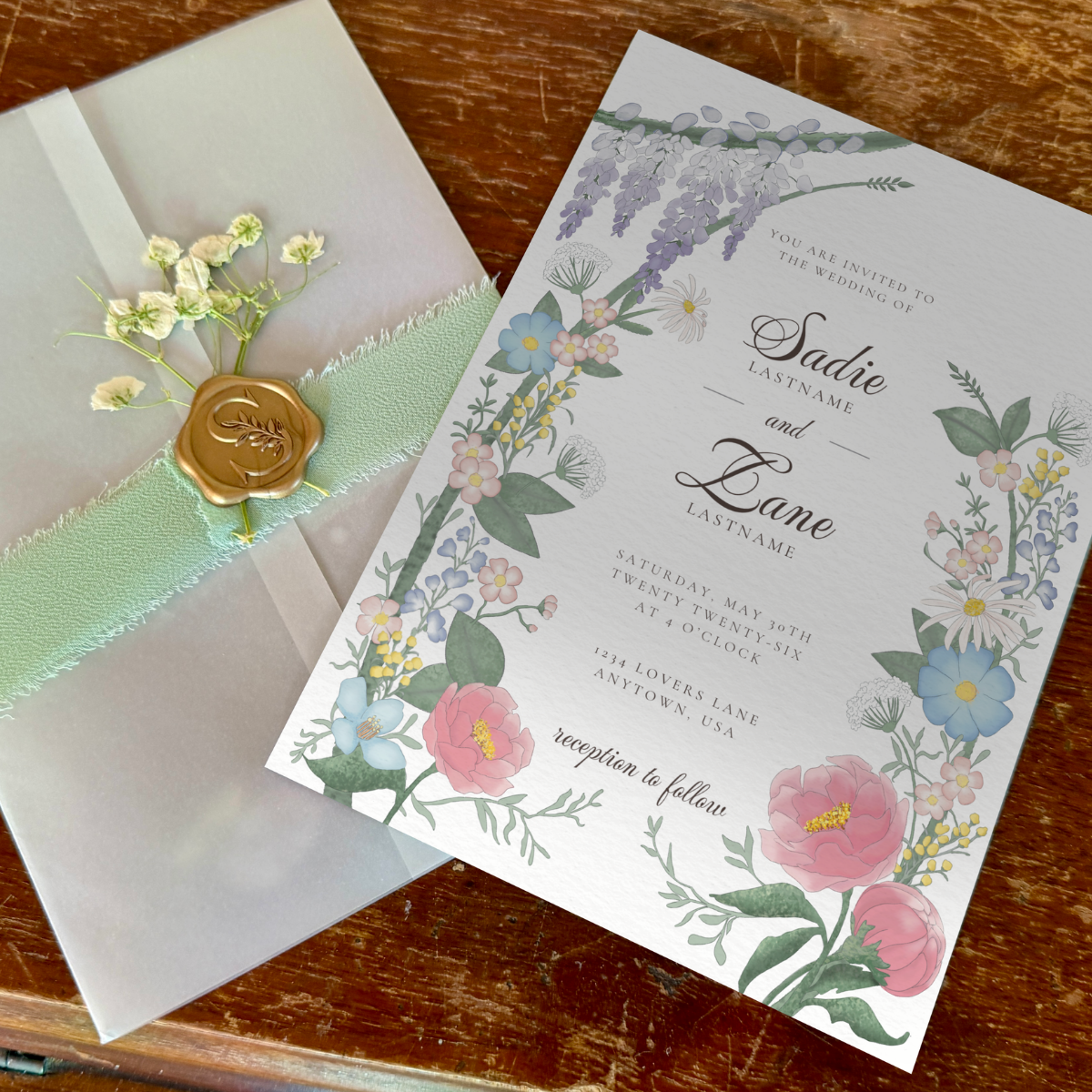 A floral wedding invitation card with pink, blue, and purple flowers arranged around elegant black script text, displayed diagonally on a rustic wooden table next to a white envelope with a mint green ribbon and gold wax seal.