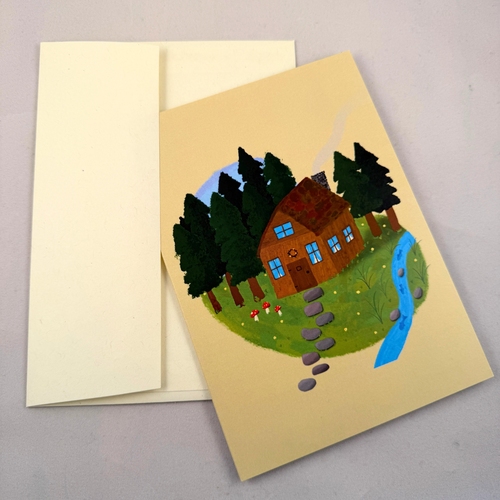A greeting card with a whimsical illustration of a brown cabin, green trees, a blue stream, and a stone path, shown with a cream envelope on a neutral gray background.