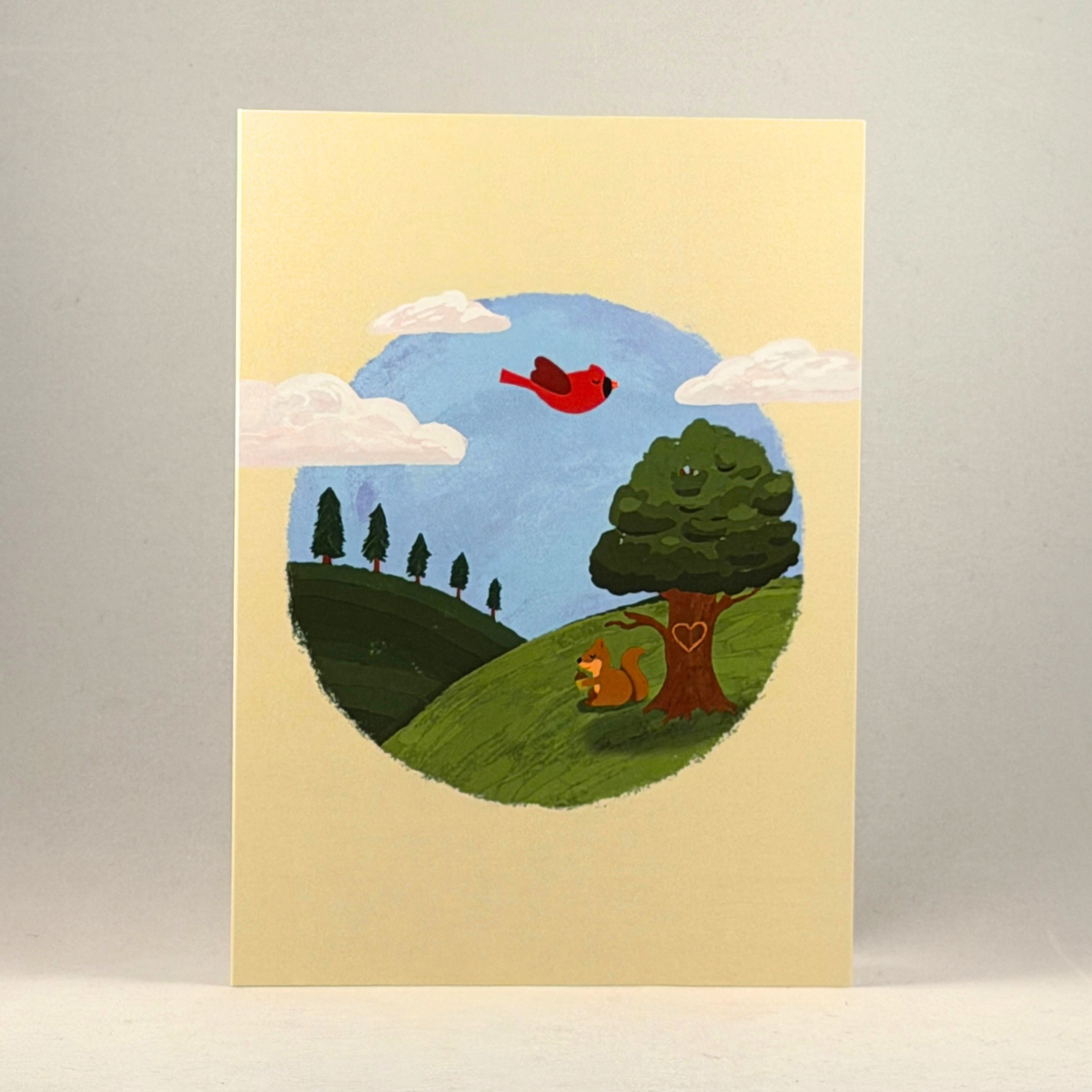 A 5x7 cream-colored greeting card featuring a hand-painted style illustration of a pastoral landscape. The scene shows a blue sky with white clouds, a red bird flying, a green hill, a tree with a heart on the trunk, and a small squirrel.
