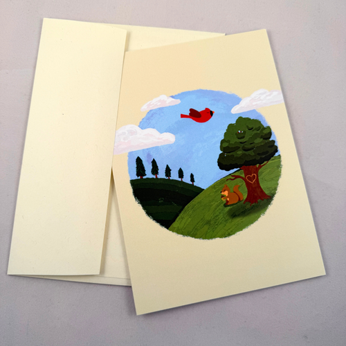A cream-colored greeting card with a whimsical painted landscape illustration, shown with its matching cream envelope on a neutral gray background. The illustration features a blue sky, green hills, a red bird, a squirrel, and a tree with a heart on its trunk.