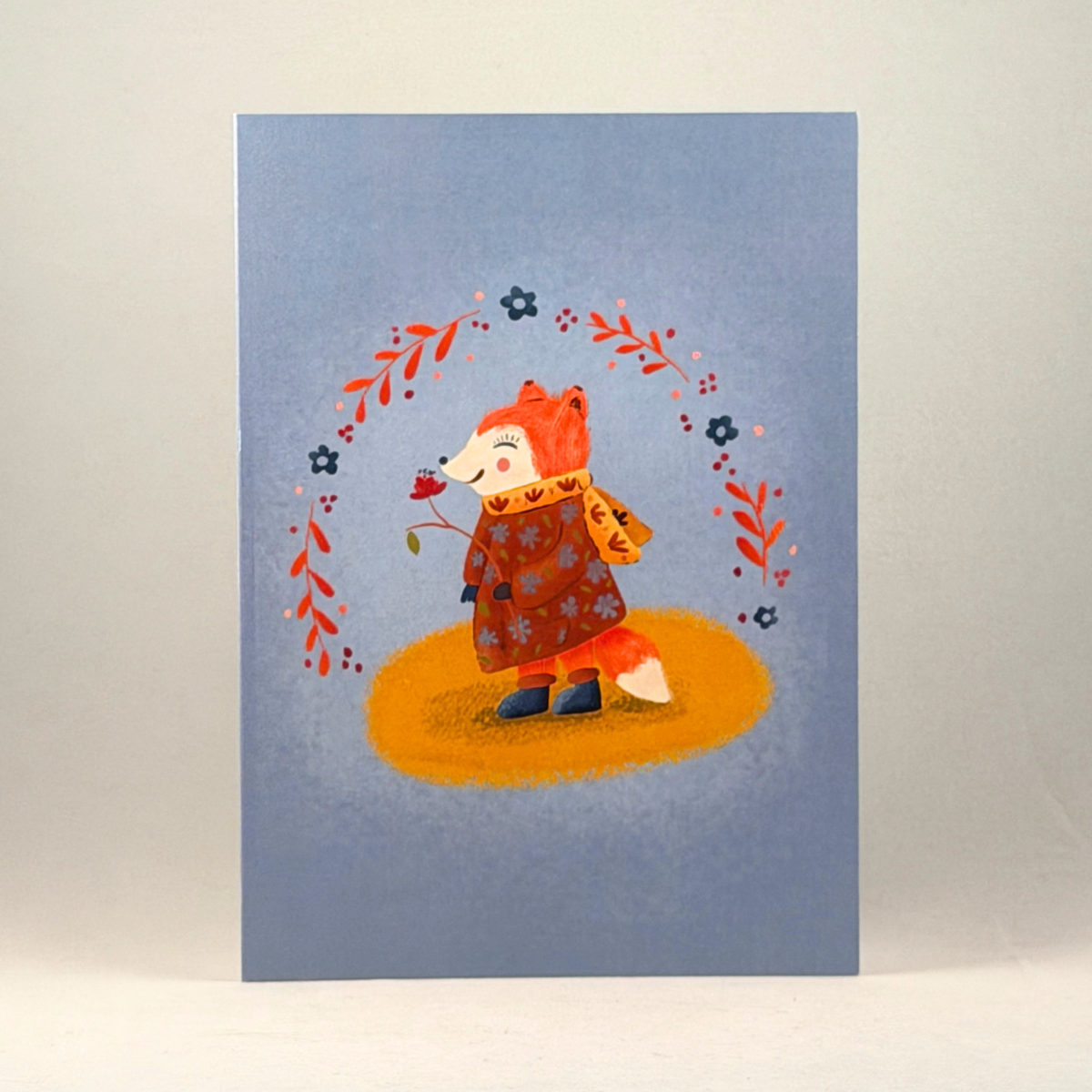 A 5x7 greeting card featuring a whimsical illustration of a fox in an autumn coat and scarf, standing on a yellow patch of ground under a circular wreath of leaves, set against a dusty blue background.