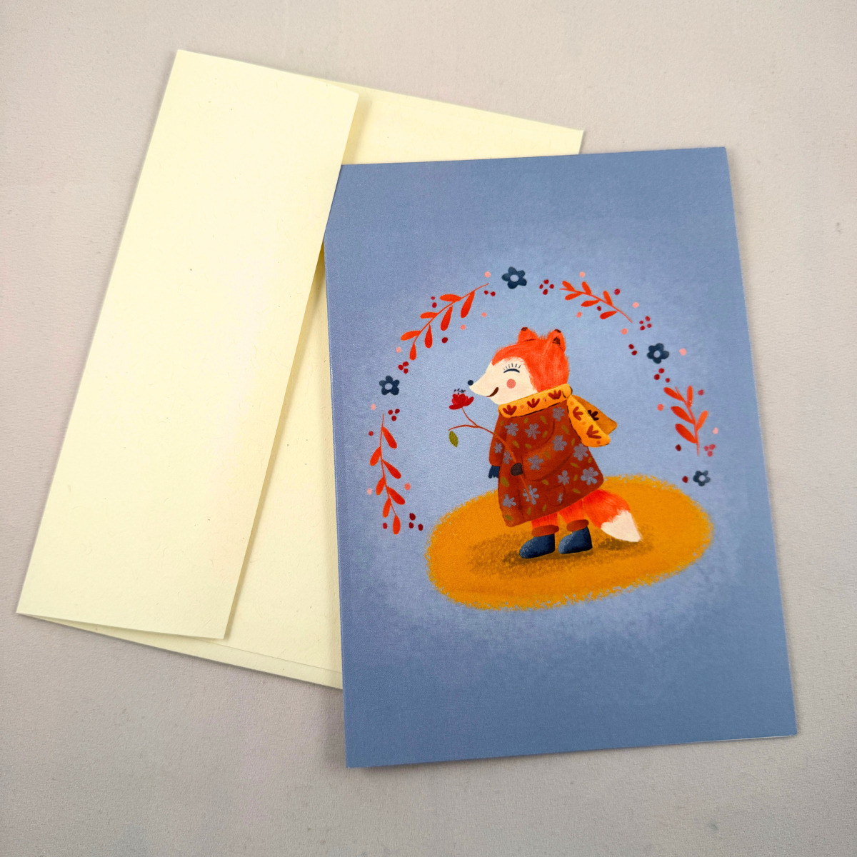 A greeting card with a whimsical illustration of a fox in a brown patterned coat standing on a yellow patch, surrounded by a floral arch, shown with a cream envelope on a neutral gray background.