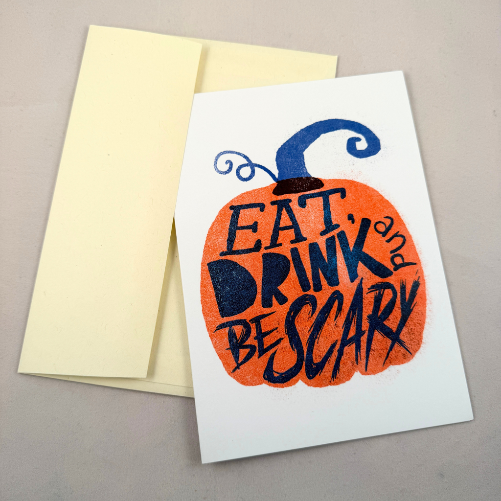 A white greeting card with a printed orange pumpkin and the phrase “EAT, DRINK, and BE SCARY” in dark blue lettering, shown with a cream envelope on a neutral gray background.