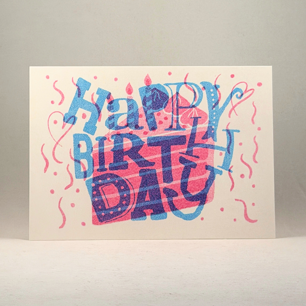 A 5x7 birthday card with a cream background featuring a colorful hand-lettered 'Happy Birthday' design in blue and pink, standing upright against a neutral gray background.