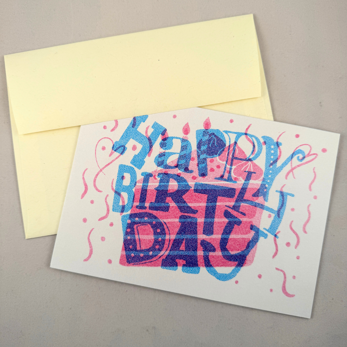 A 5x7 birthday card with a cream envelope, featuring a colorful hand-lettered 'Happy Birthday' design in blue and pink over a pink cake illustration, photographed on a neutral gray background.