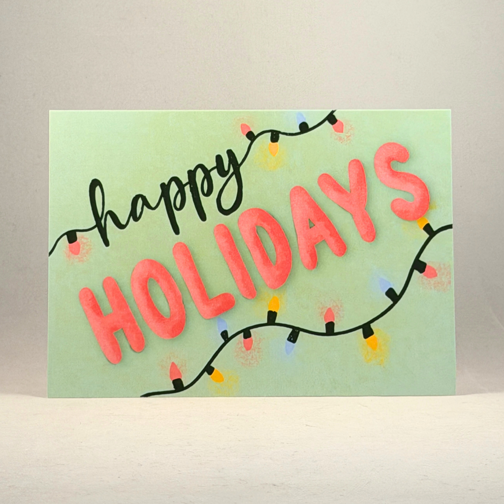 A 5x7 holiday greeting card with 'happy HOLIDAYS' written in black and coral pink letters, decorated with string lights on a light green background, set against a neutral gray backdrop.