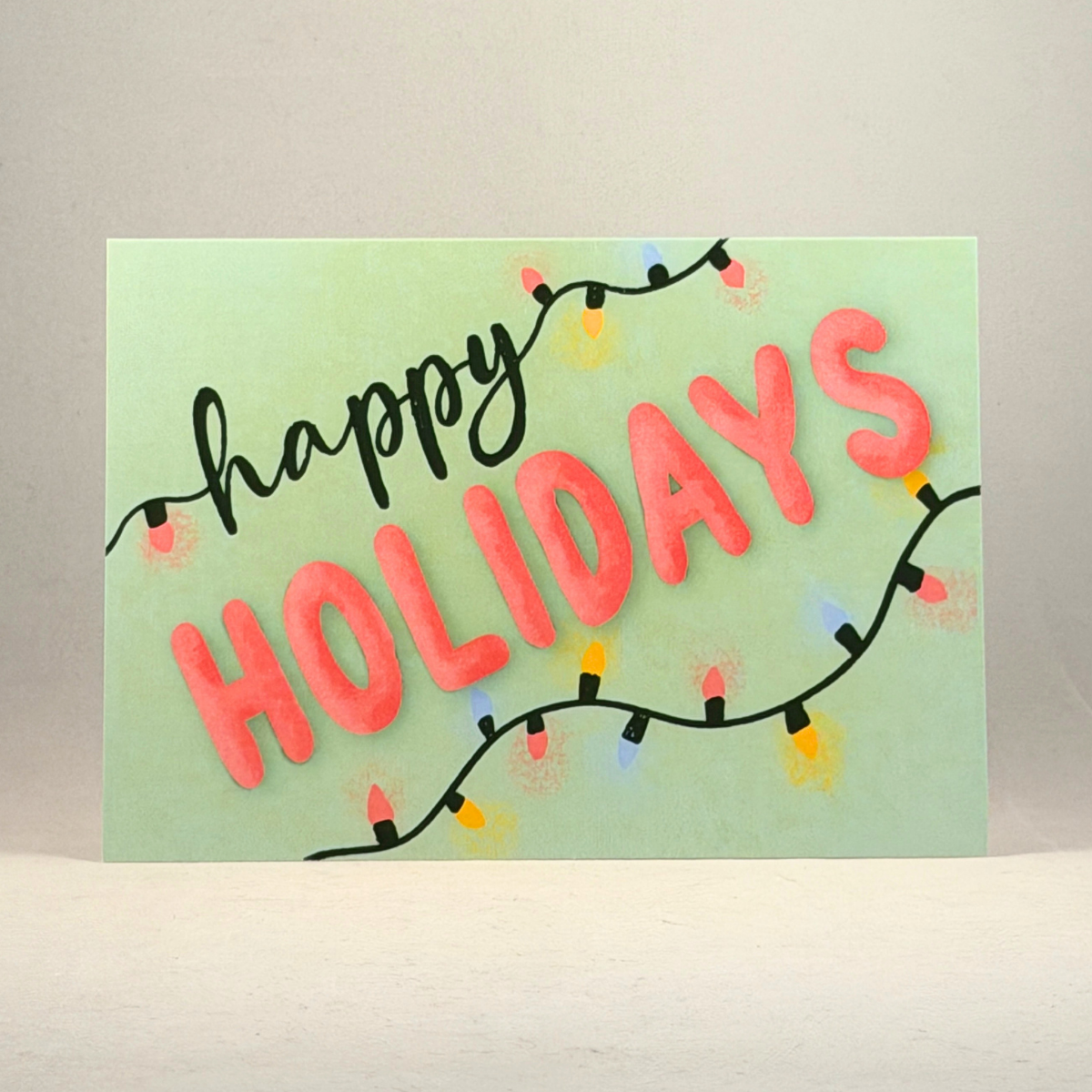 A 5x7 holiday greeting card with 'happy HOLIDAYS' written in black and coral pink letters, decorated with string lights on a light green background, set against a neutral gray backdrop.