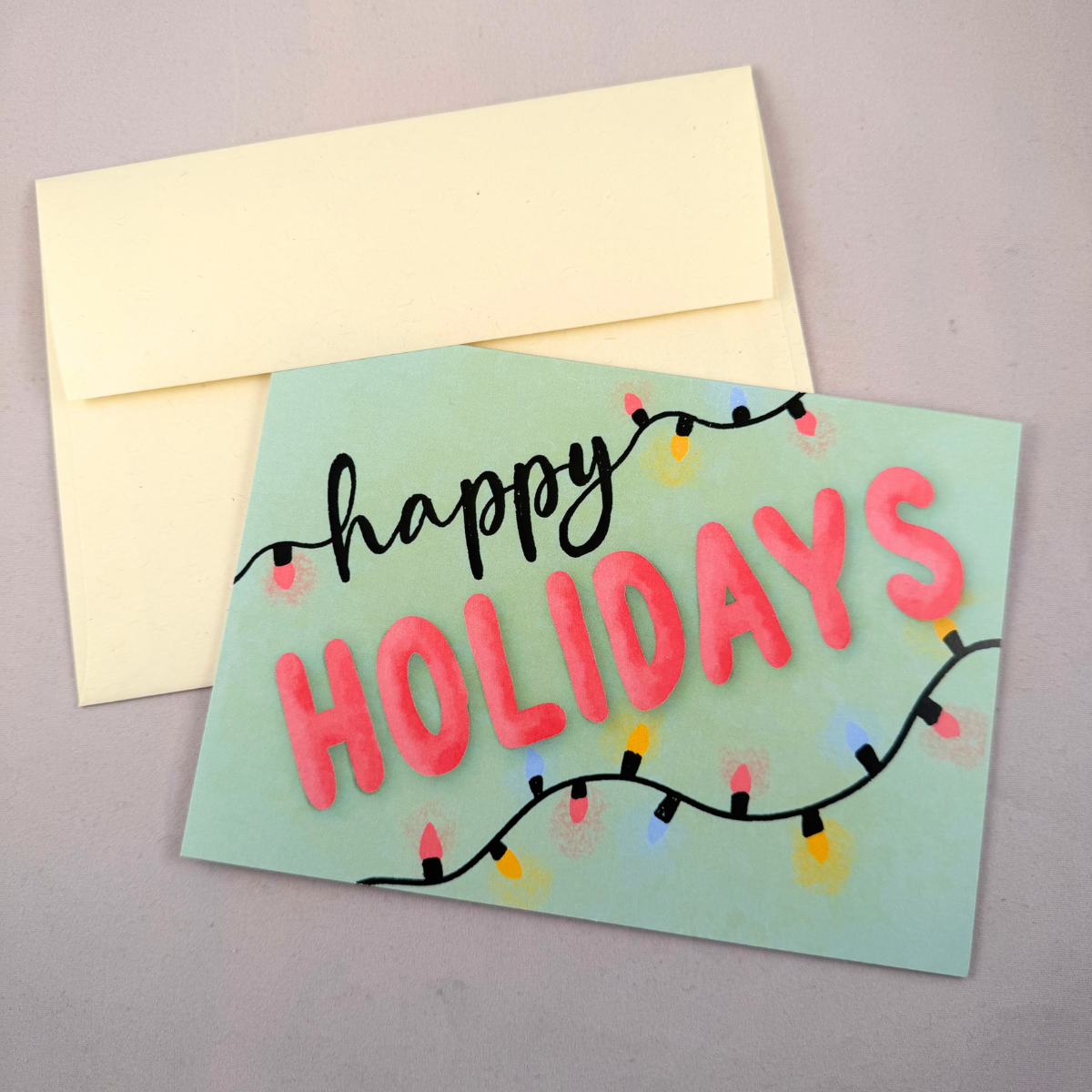 A holiday greeting card with a pale mint green background, featuring the words 'happy HOLIDAYS' in black and pink lettering, decorated with hand-painted string lights, shown with a cream-colored envelope on a neutral gray background.
