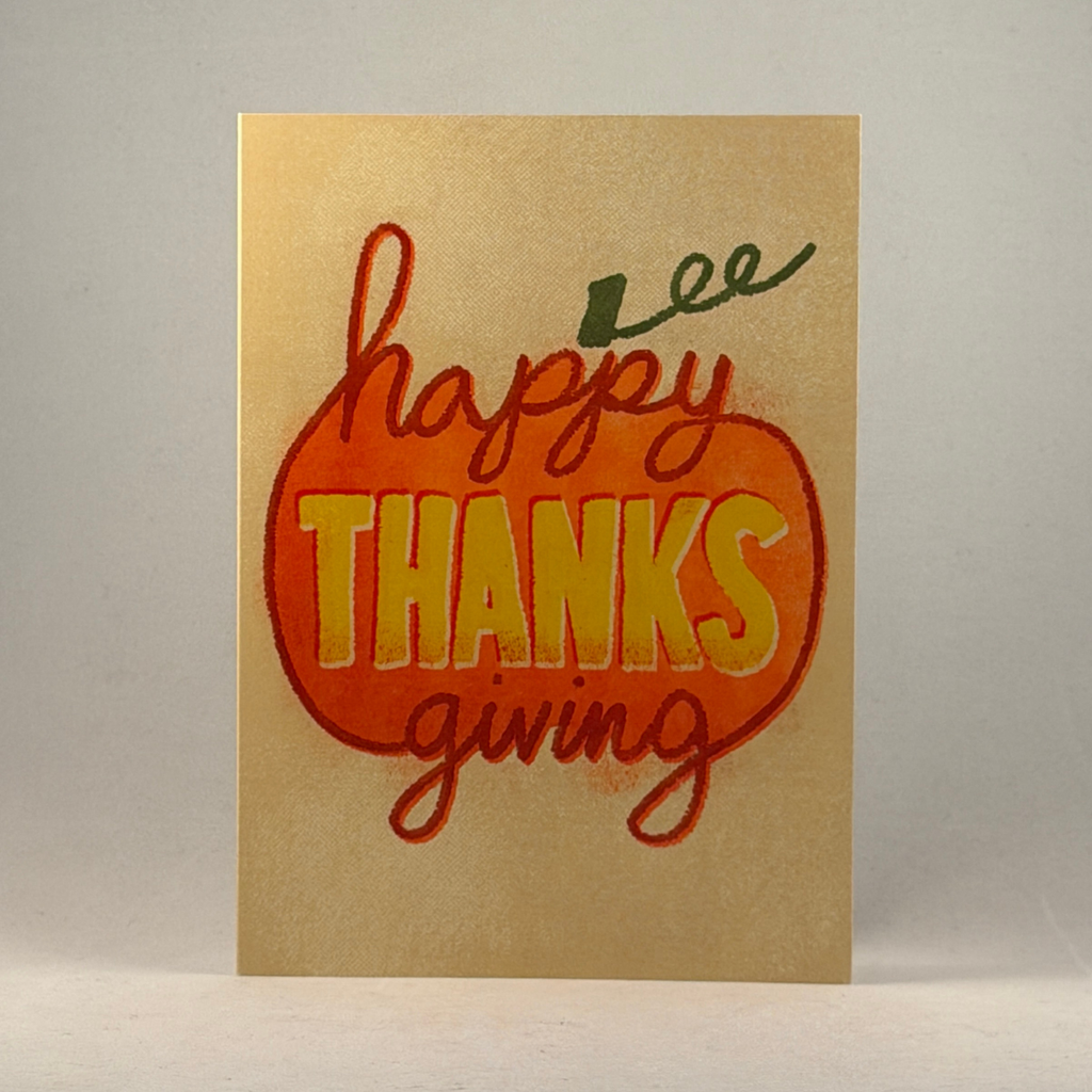 A 5x7 greeting card with a metallic gold background, featuring a large orange pumpkin outline and the words 'happy THANKS giving' in red and yellow lettering, photographed against a neutral gray background.