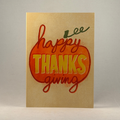 A 5x7 greeting card with a metallic gold background, featuring a large orange pumpkin outline and the words 'happy THANKS giving' in red and yellow lettering, photographed against a neutral gray background.