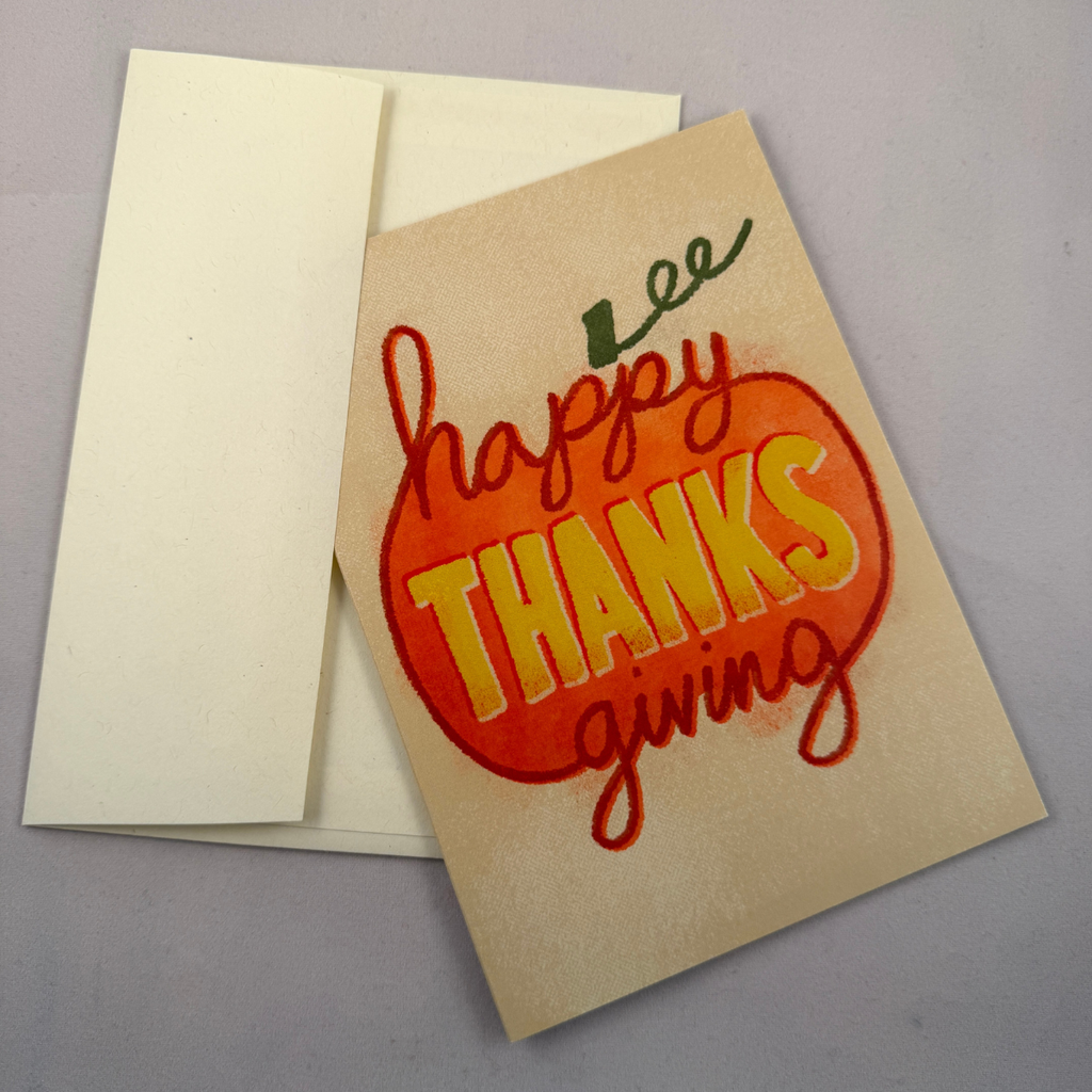A Thanksgiving greeting card with a cream envelope, featuring a hand-painted orange pumpkin design with the words 'happy THANKS giving' in red and yellow lettering.
