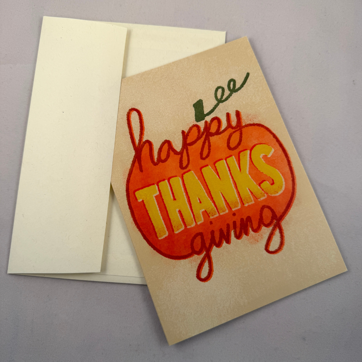 A Thanksgiving greeting card with a cream envelope, featuring a hand-painted orange pumpkin design with the words 'happy THANKS giving' in red and yellow lettering.