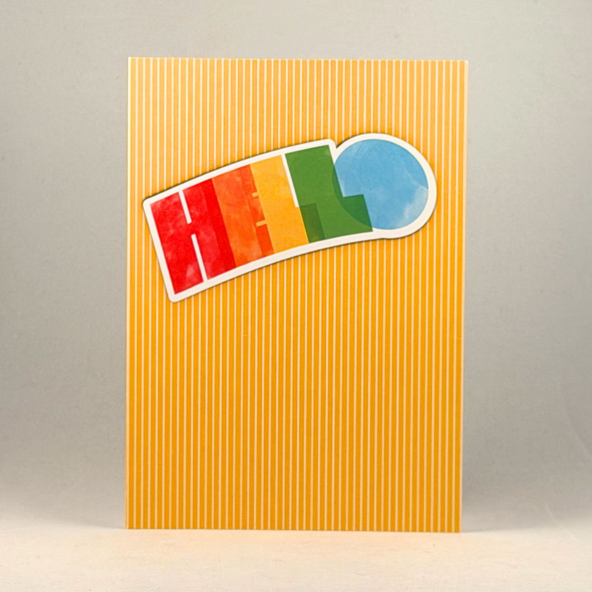 A 5x7 greeting card with a yellow and white vertical striped background, featuring a bright 'HELLO' in rainbow colors.