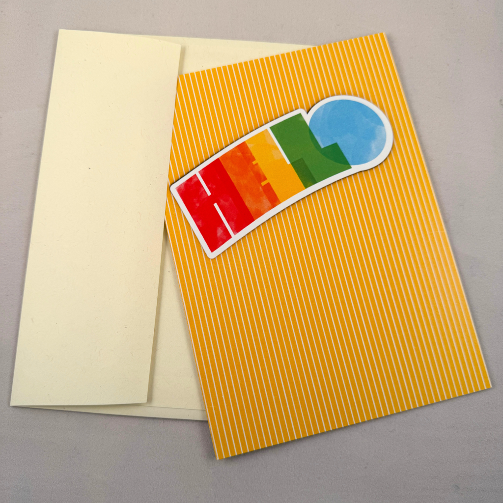 A greeting card with a bright yellow and white striped pattern, featuring a colorful 'HELLO' design in rainbow colors and a blue circle, shown with a cream-colored envelope on a neutral gray background.