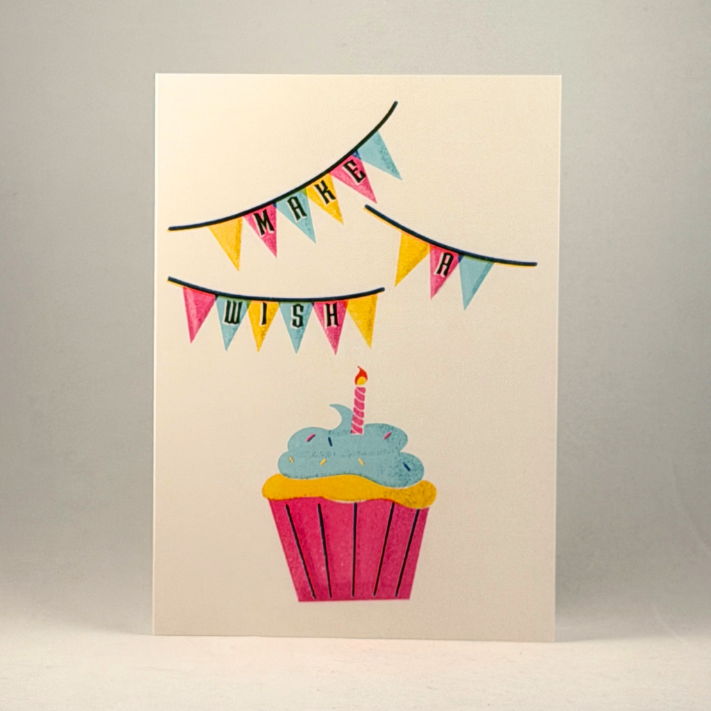 A 5x7 greeting card with a colorful illustration of a cupcake and bunting that reads 'MAKE A WISH', photographed against a neutral gray background.