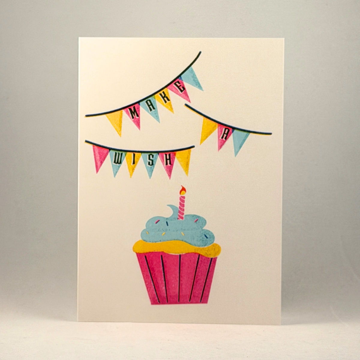 A 5x7 greeting card with a colorful illustration of a cupcake and bunting that reads 'MAKE A WISH', photographed against a neutral gray background.