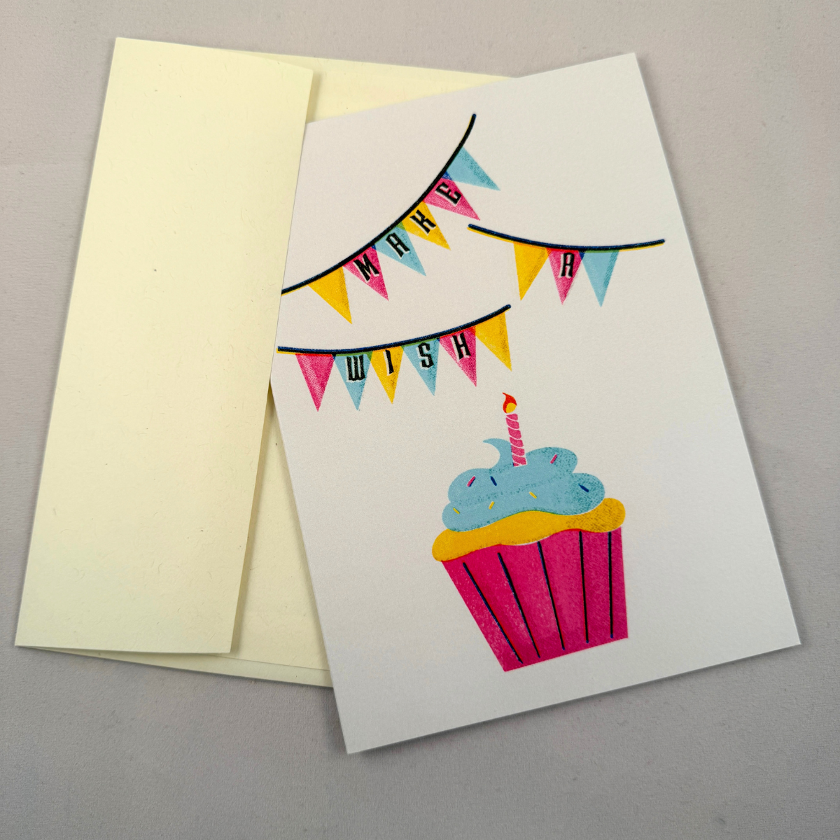 A white birthday card with a colorful illustration of a cupcake and bunting banner reading 'MAKE A WISH' is placed on top of a cream envelope against a neutral gray background.