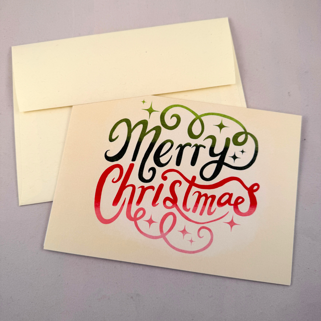 A cream-colored Christmas greeting card with 'Merry Christmas' written in black and red script, with a matching cream envelope behind it on a gray background.
