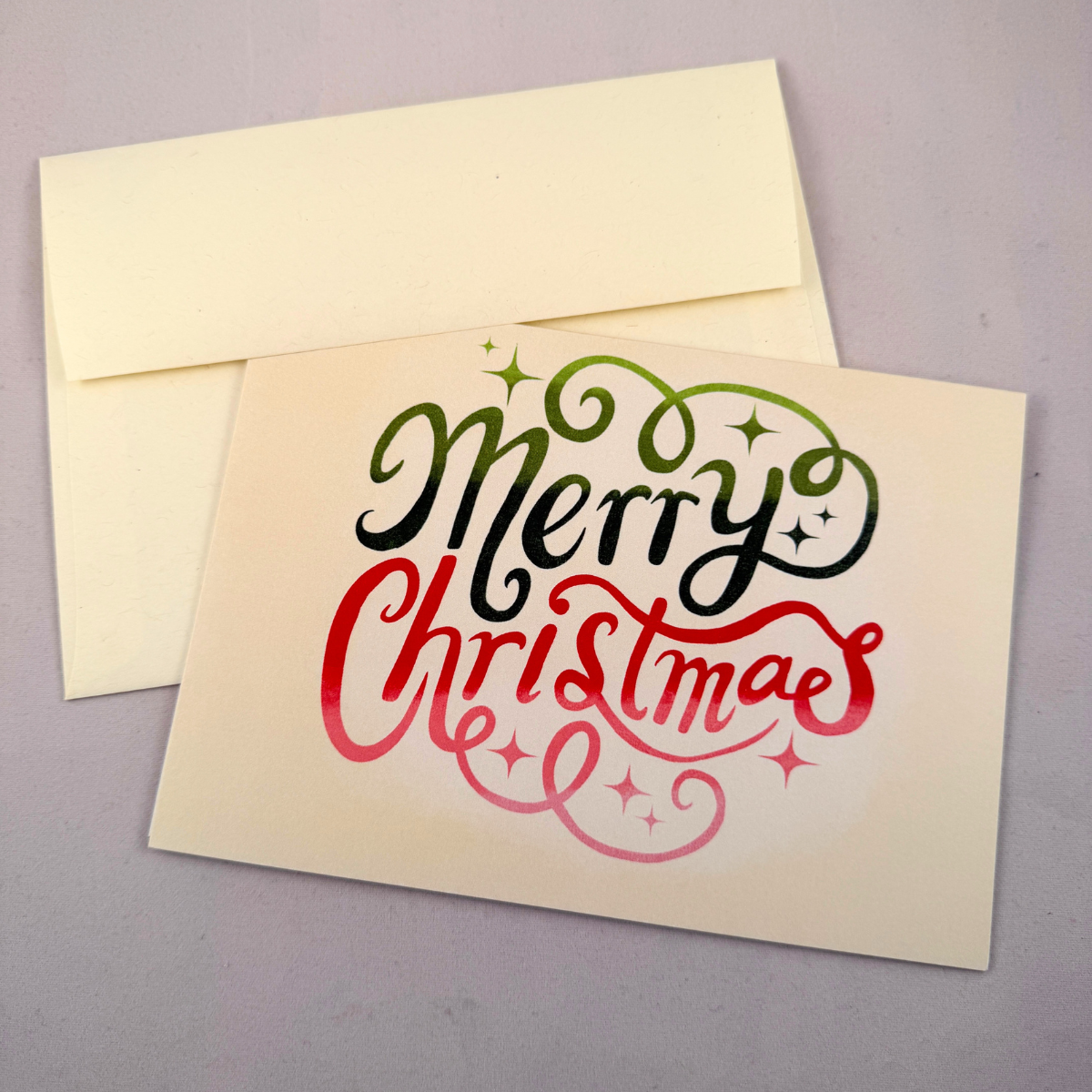 A cream-colored Christmas greeting card with 'Merry Christmas' written in black and red script, with a matching cream envelope behind it on a gray background.