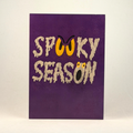 A 5x7 purple greeting card featuring the words 'SPOOKY SEASON' in a dripping beige font, with a yellow sinister face incorporated into the lettering, photographed against a neutral gray background.