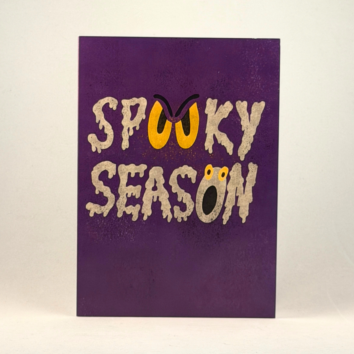 A 5x7 purple greeting card featuring the words 'SPOOKY SEASON' in a dripping beige font, with a yellow sinister face incorporated into the lettering, photographed against a neutral gray background.