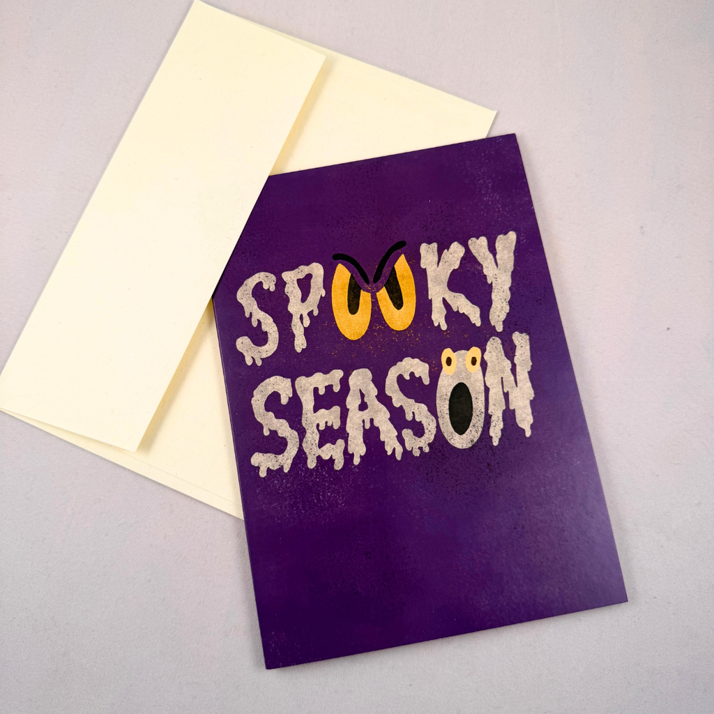 A purple Halloween greeting card with the words 'SPOOKY SEASON' in white dripping letters, featuring a yellow sinister eyes as the 'OO' in the word 'spooky' and a shocked mouth as the last 'O' in the word 'season', shown with a cream envelope on a light gray background.