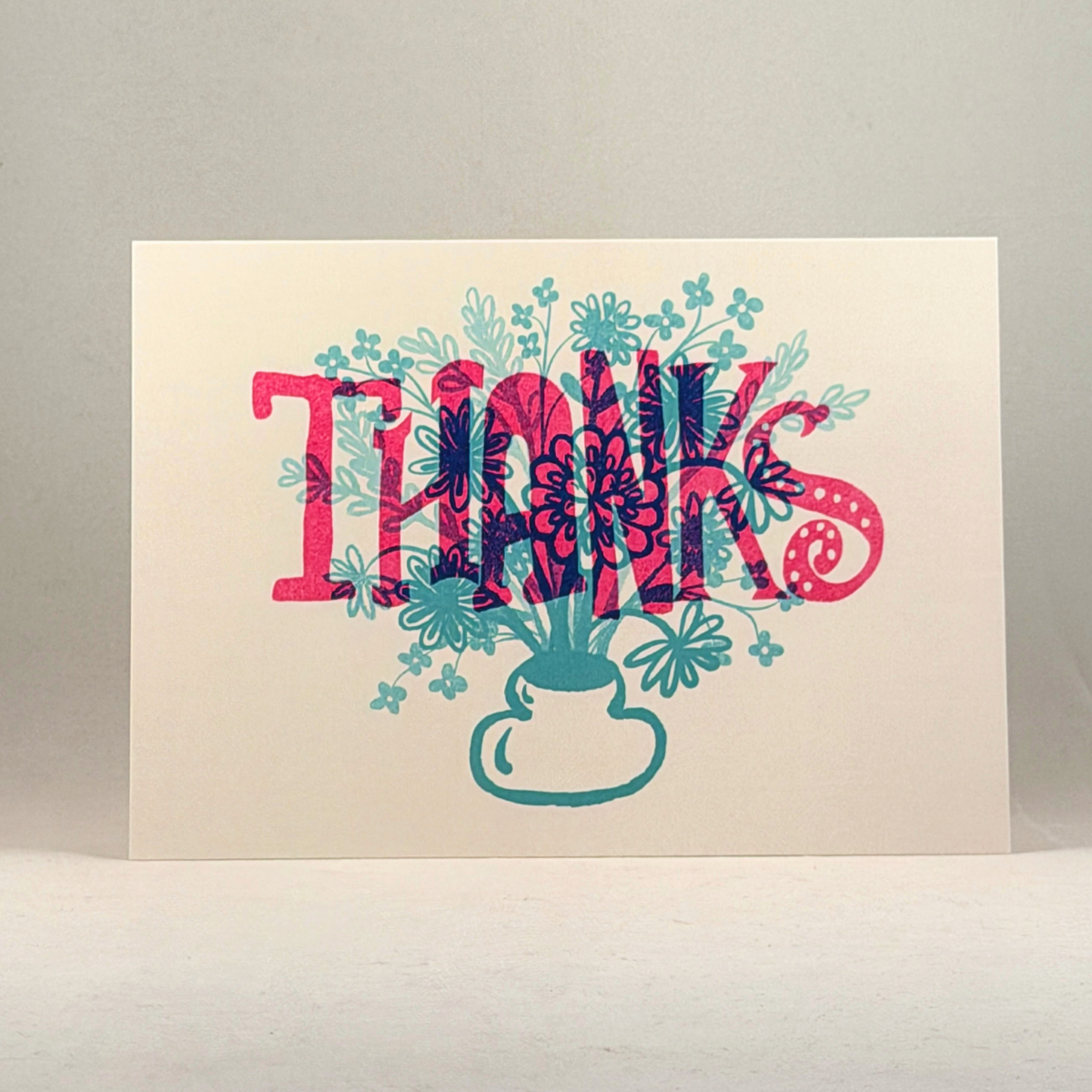 A 5x7 white greeting card featuring the word 'THANKS' in large pink letters with teal floral and botanical details layered behind and around it, plus a teal vase illustration at the bottom, photographed against a neutral gray background.