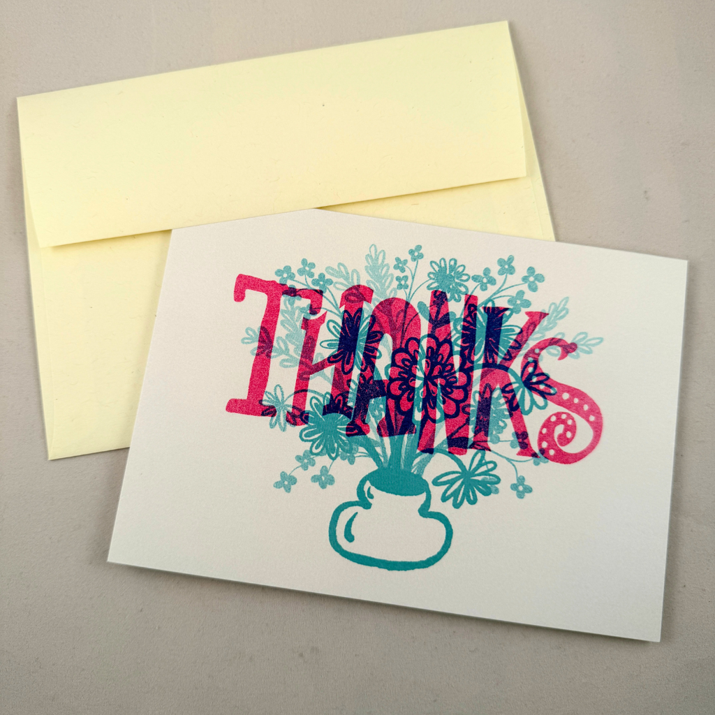 A white greeting card with a colorful floral 'THANKS' design in red, pink, and teal, shown with a cream envelope on a neutral gray background.