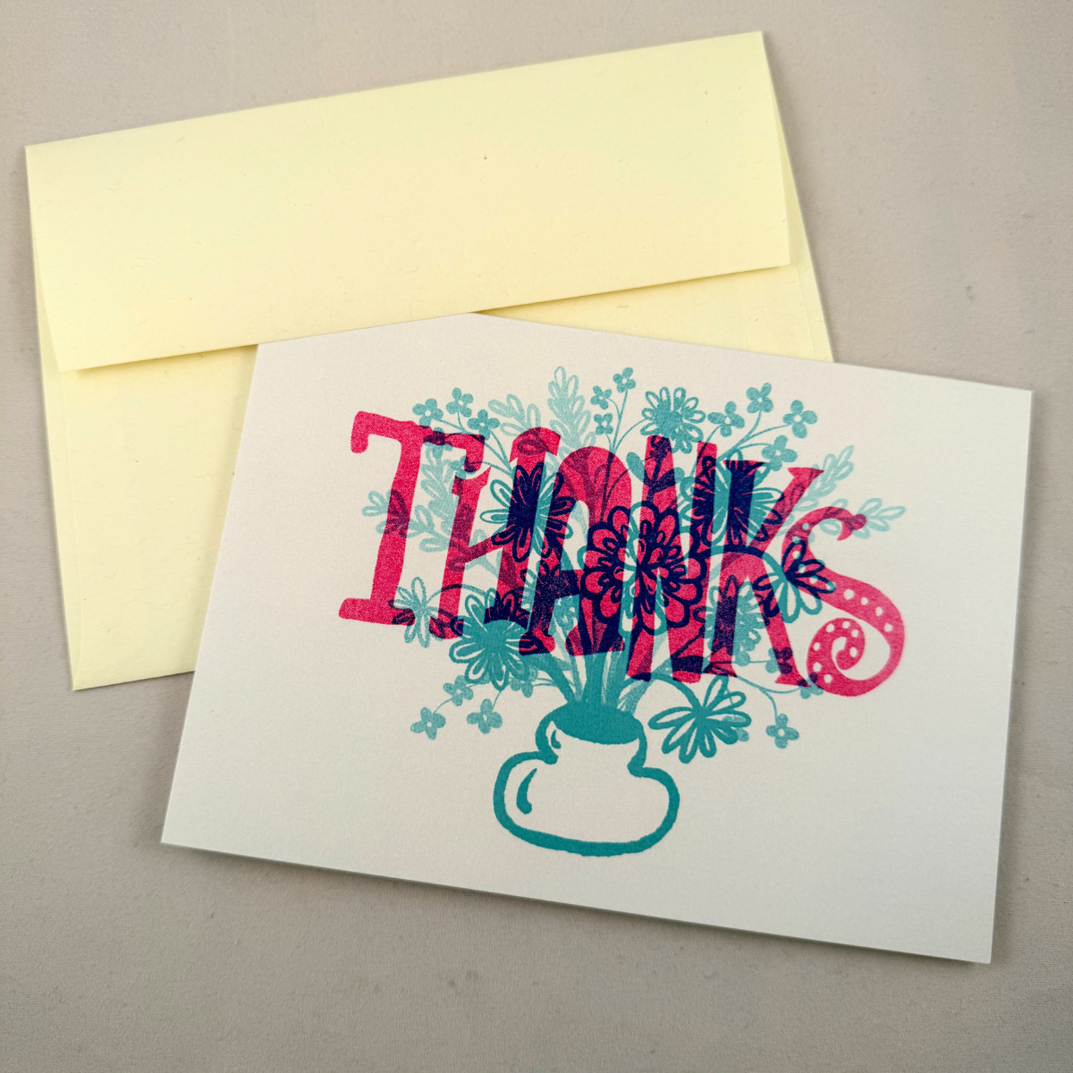 A white greeting card with a colorful floral 'THANKS' design in red, pink, and teal, shown with a cream envelope on a neutral gray background.