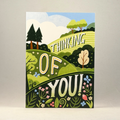 A 5x7 greeting card featuring a colorful landscape illustration with the words 'THINKING OF YOU!' in large white letters. The design includes green hills, trees, flowers, and a blue sky, photographed against a plain neutral gray background.