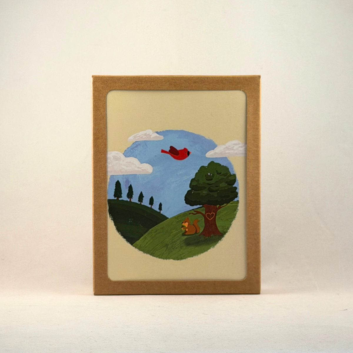 A boxed notecard set with a windowed kraft brown box, featuring a cream-colored greeting card with a hand-painted circular landscape illustration, set against the background near the top of the card. 