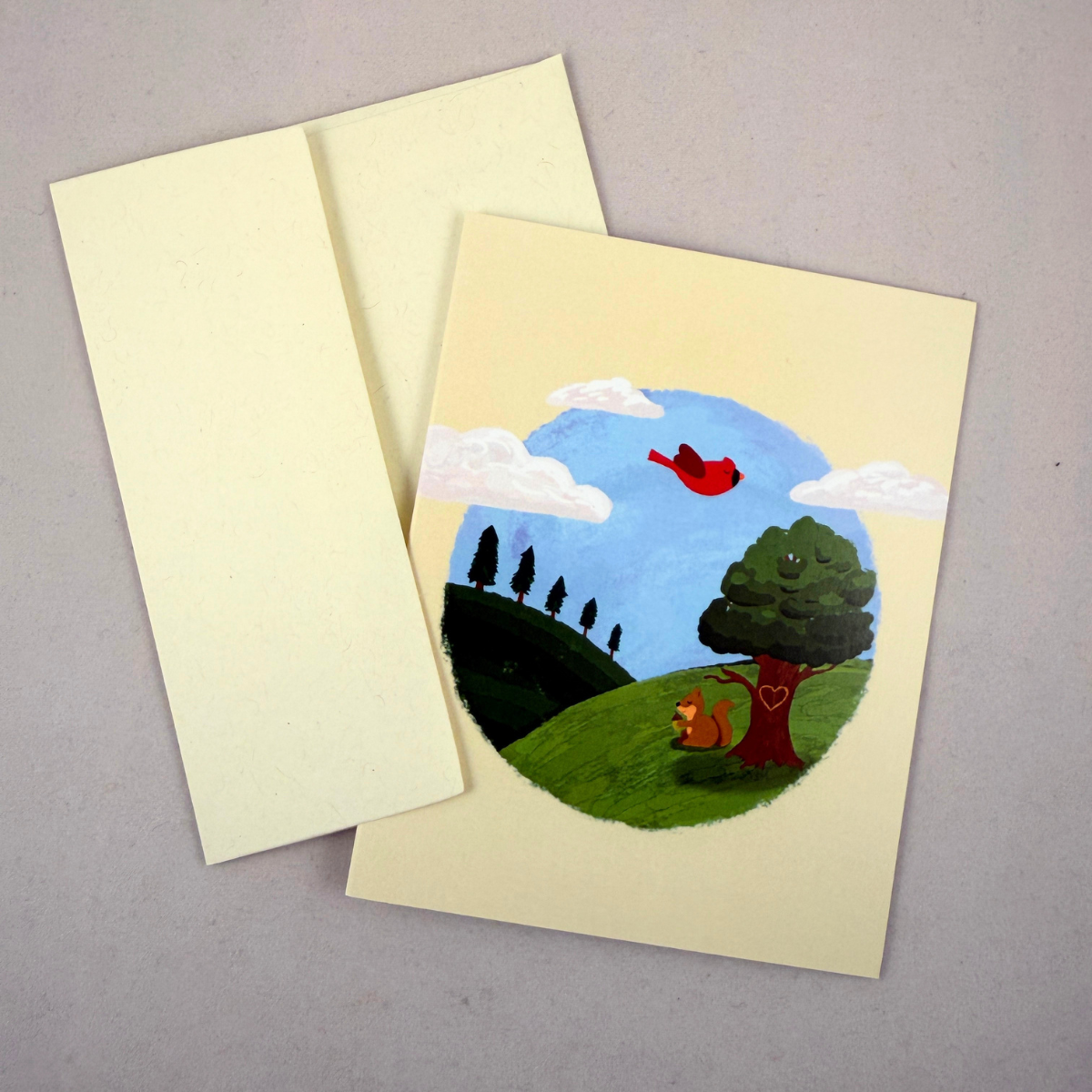 A cream-colored notecard with a hand-painted circular landscape illustration, shown with its matching cream envelope on a neutral gray background.