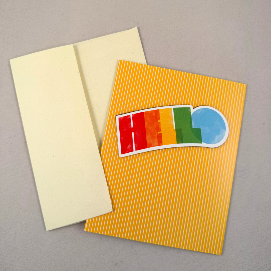 A notecard with thin yellow vertical stripes and colorful 'HELLO' lettering on the front, shown with a cream envelope on a neutral gray background.
