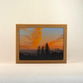 A kraft paper notecard box with an open window displaying a sunset landscape print of orange clouds and dark pine tree silhouettes, set against a plain off-white background.