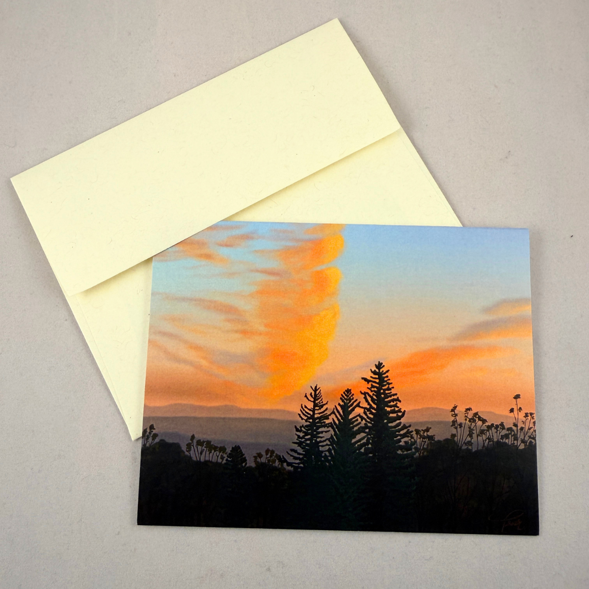 A notecard showing a sunset landscape with orange and blue sky, clouds, mountains, and dark tree silhouettes, placed on a light gray surface with a cream envelope behind it.