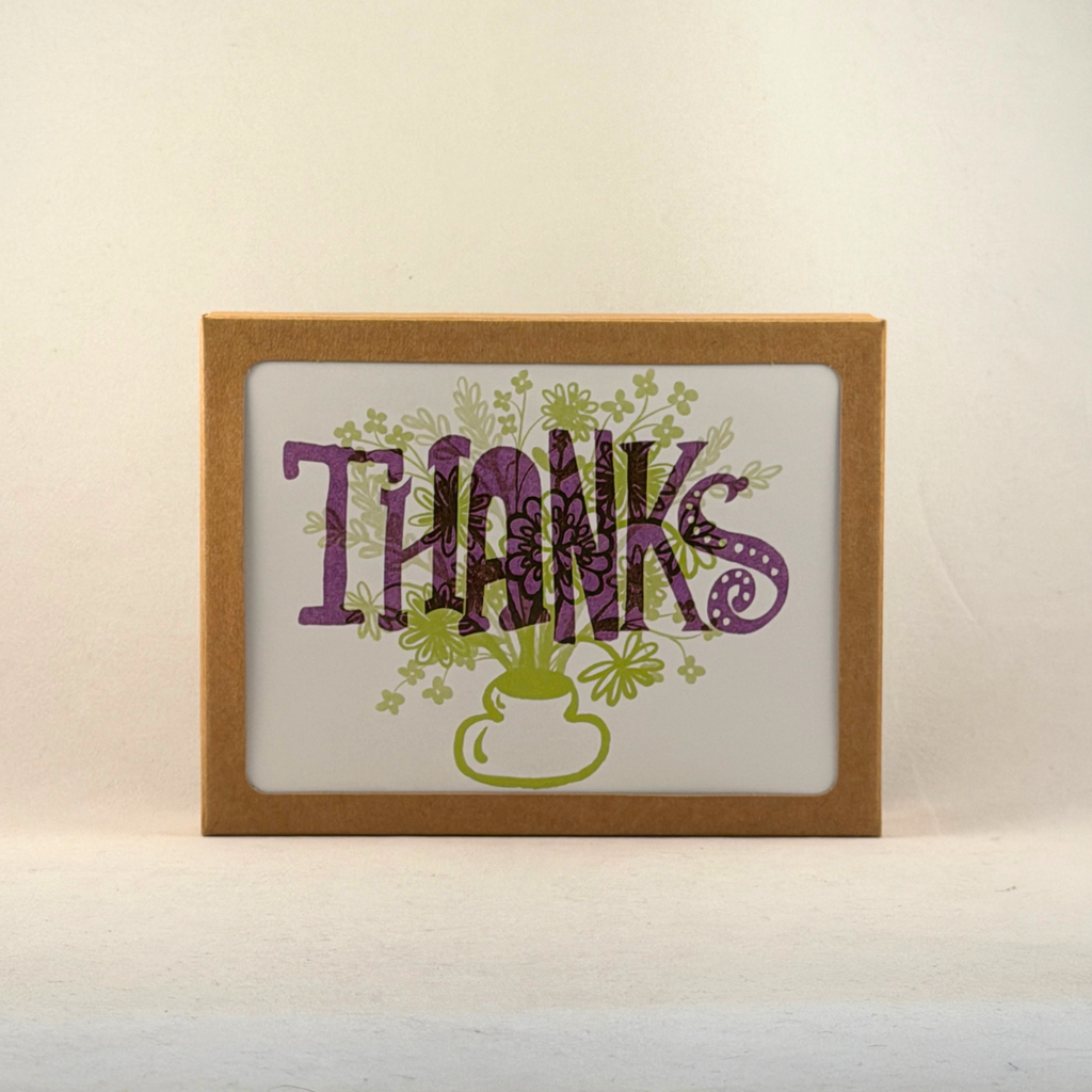 A boxed notecard set with a windowed kraft brown box, featuring the word 'THANKS' in purple floral lettering with green flowers and a green pot illustration on a white background.