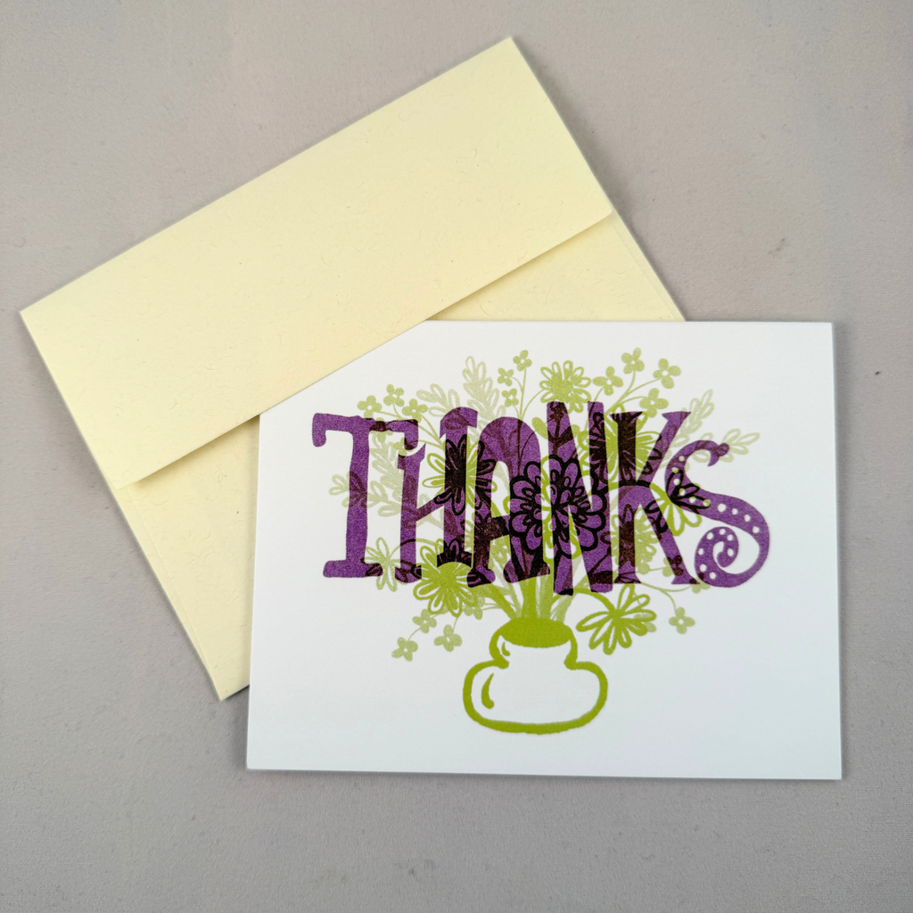 A white greeting card with the word 'THANKS' in large purple letters, decorated with green floral and clover motifs, shown with a cream envelope on a neutral gray background.
