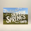 A postcard featuring a scenic landscape of green hills under a blue sky with white clouds, overlaid with large white decorative text reading 'Greetings from SHARON SPRINGS NEW YORK.'