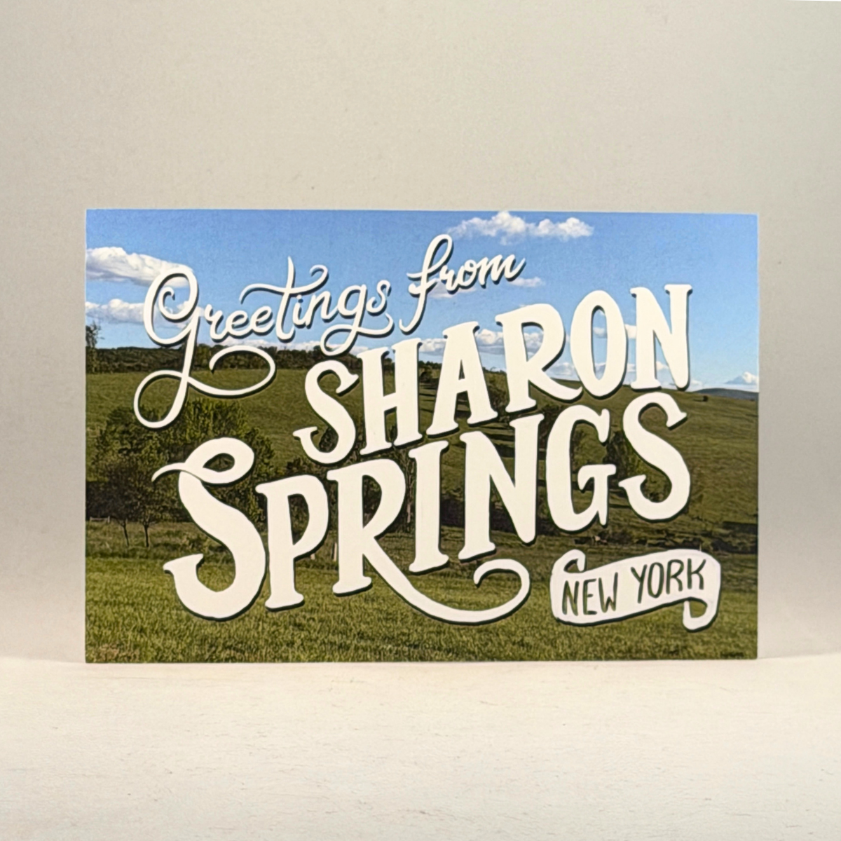 A postcard featuring a scenic landscape of green hills under a blue sky with white clouds, overlaid with large white decorative text reading 'Greetings from SHARON SPRINGS NEW YORK.'