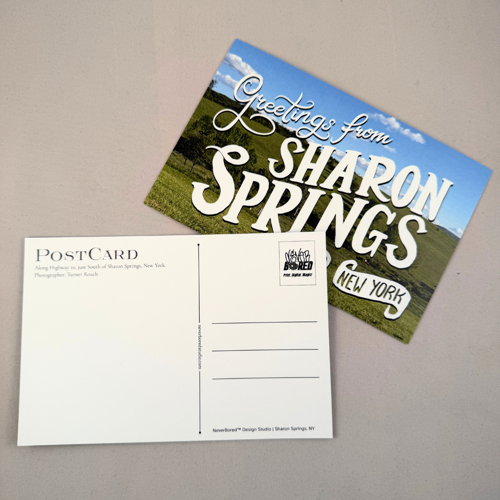 A postcard set on a neutral gray surface, showing both the front and back. The front features a scenic landscape of green fields under a blue sky with the text 'Greetings from SHARON SPRINGS NEW YORK'.
