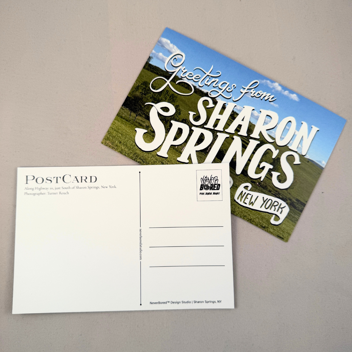 A postcard set on a neutral gray surface, showing both the front and back. The front features a scenic landscape of green fields under a blue sky with the text 'Greetings from SHARON SPRINGS NEW YORK'.