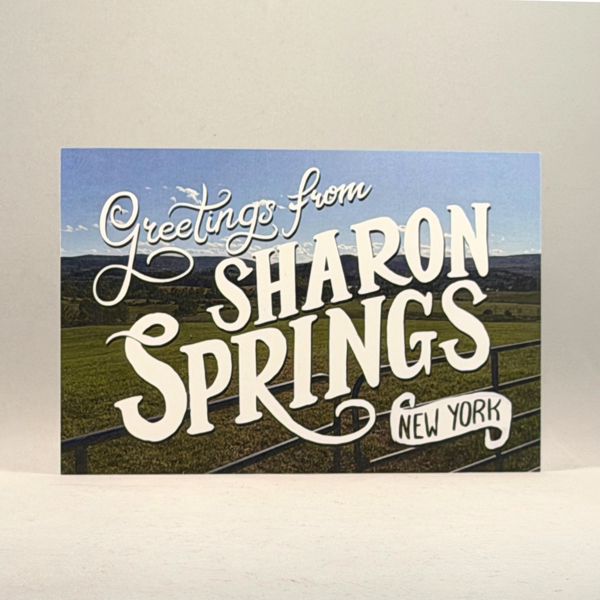 A postcard featuring a scenic landscape of Sharon Springs, New York, with large white decorative text reading 'Greetings from SHARON SPRINGS NEW YORK' over a blue sky and green valley scene.