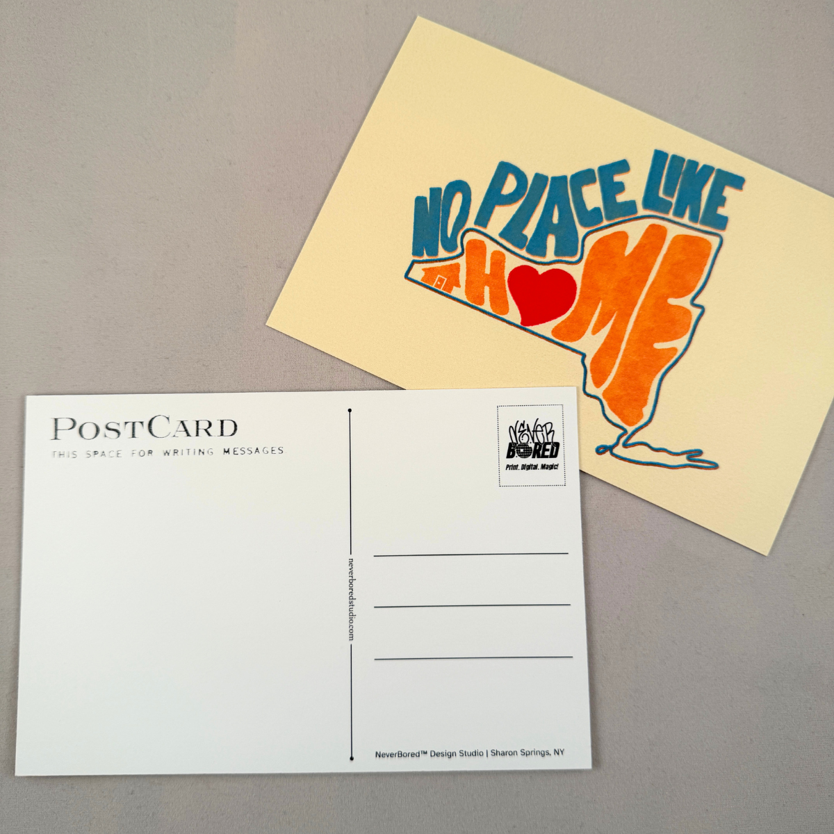 A postcard set on a gray surface, including the back of a postcard with printed text lines and a cream postcard featuring a colorful graphic of the state of New York in orange and blue with a red heart and the words 'NO PLACE LIKE HOME'.