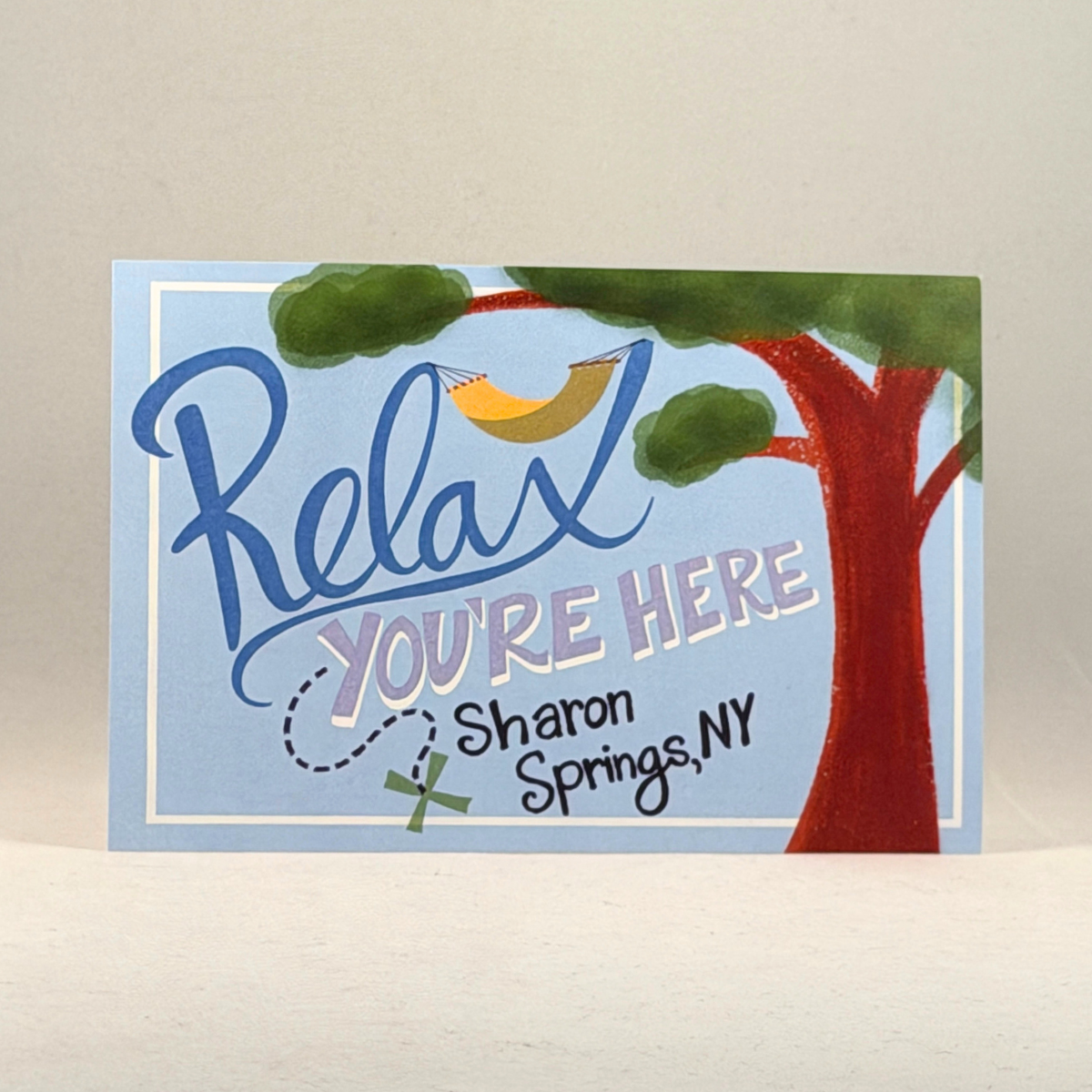A postcard with a painted tree and the words 'Relax YOU'RE HERE Sharon Springs, NY' on a light blue background, photographed against a neutral gray background.