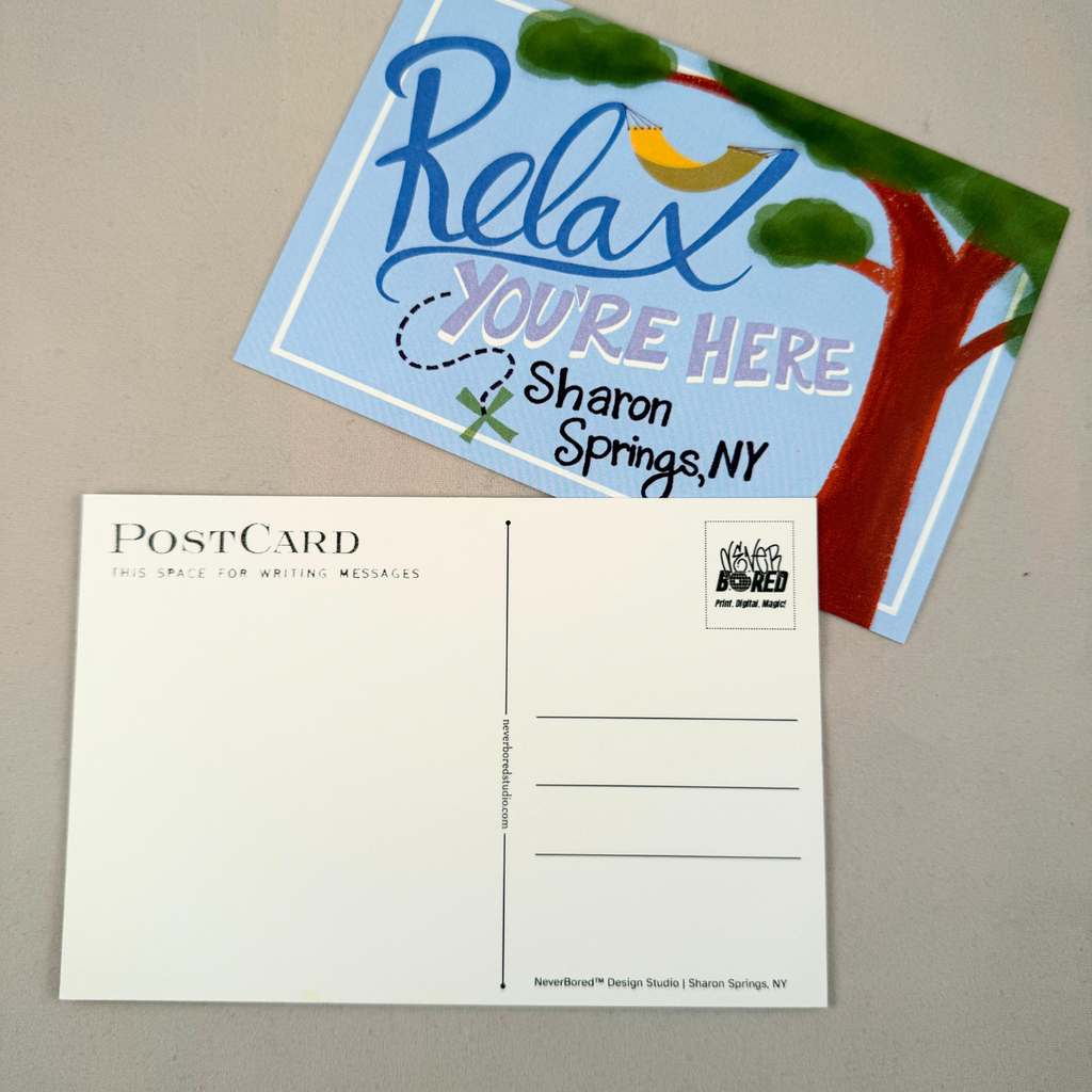 A postcard set on a neutral gray surface, featuring a white back side with printed postcard lines and a colorful front side showing a blue 'Relax YOU'RE HERE' design with a tree and the text 'Sharon Springs, NY'.